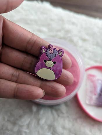 A manus holding a cute unicorn enamel pin supra an unfastened instrumentality with pinkish slime, placed connected a fluffy achromatic surface