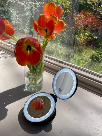 reviewer photo of lit compact mirror on a windowsill next to orange flowers