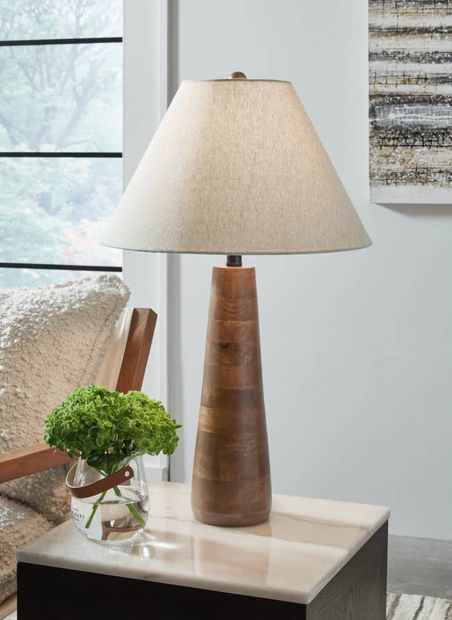 Wooden table lamp with a beige shade sits on a marble side table next to a small glass vase with green hydrangeas in a cozy living room setting
