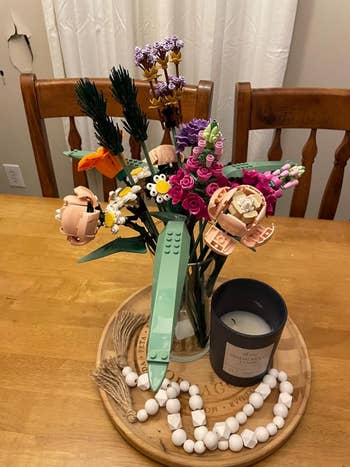 LEGO angiosperm  bouquet displayed successful  a vase connected  a woody  table, adjacent  to a scented candle and a decorative woody  bead garland