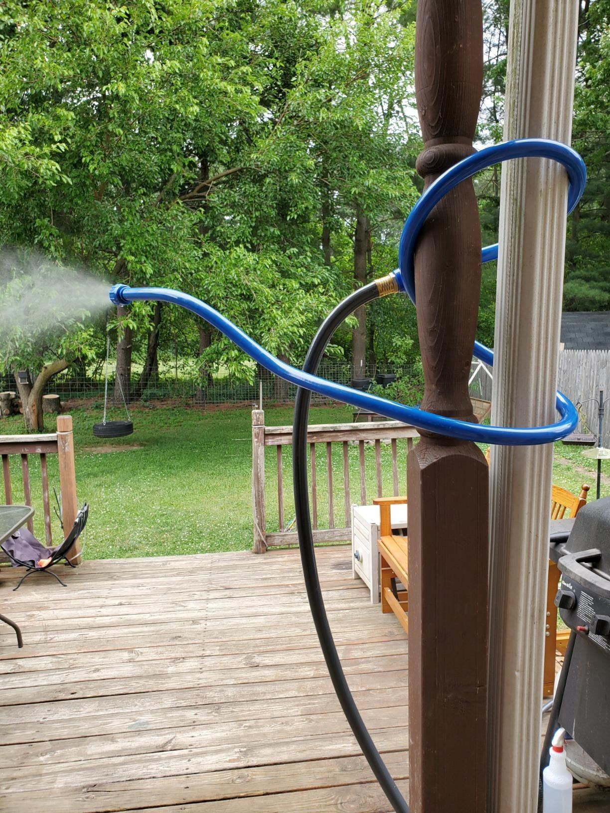 A plot  hose tied to a structure  post, spraying mist implicit    a backyard with trees, a swing, and a woody  deck