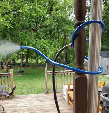 A plot hose tied to a structure post, spraying mist implicit a backyard with trees, a swing, and a woody deck
