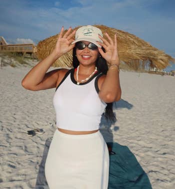 Person connected a formation wearing a achromatic vessel top, achromatic skirt, and hat, smiling and adjusting sunglasses