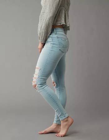 A person models light-wash, ripped skinny jeans, standing barefoot against a plain background