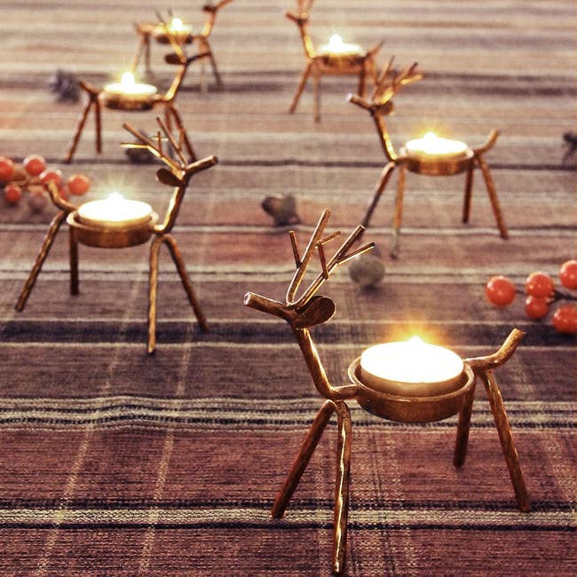 Reindeer-shaped candle holders displayed on a plaid surface, each holding a lit tealight, surrounded by decorative berries and pinecones