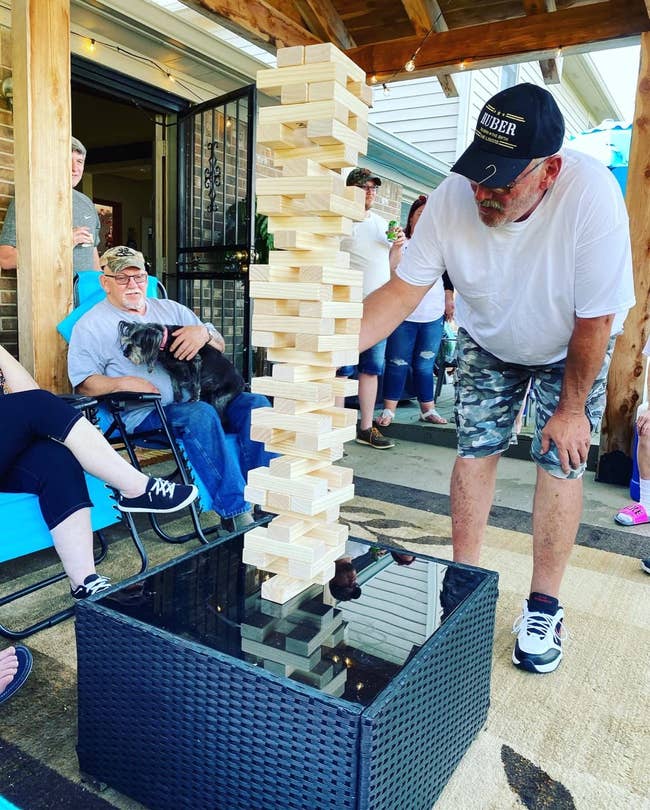 Man wearing casual apparel and chapeau playing a ample outdoor Jenga crippled connected a patio. Others beryllium nearby, with a canine connected 1 person's lap