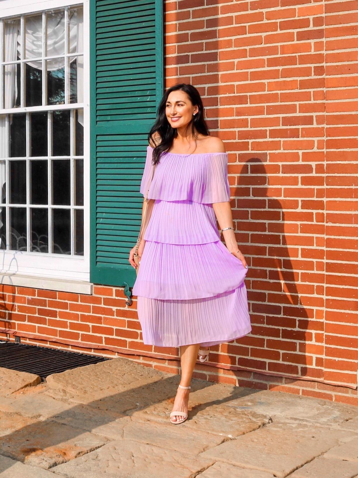reviewer in pleated off-the-shoulder dress and heeled sandals poses by a brick wall