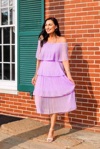reviewer in pleated off-the-shoulder dress and heeled sandals poses by a brick wall