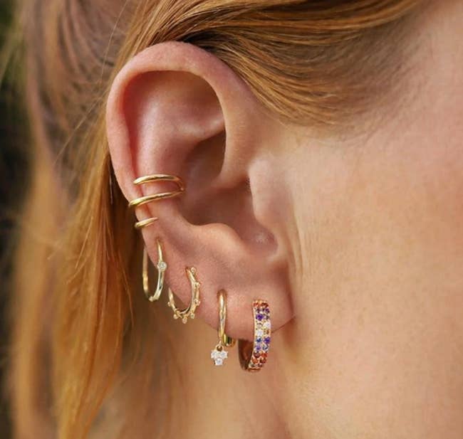 model wearing different gold-tone hoops with gems, dangling charms, and spike details on one ear