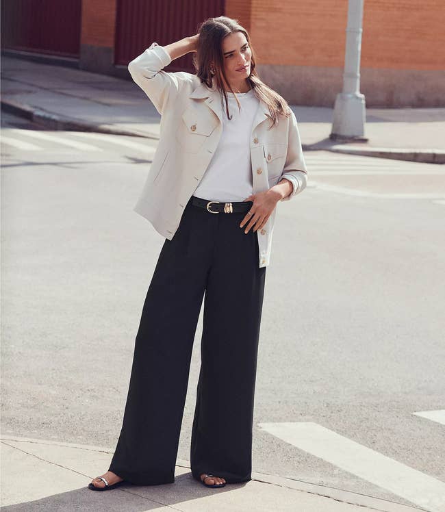 Person wearing a light jacket, white top, and wide-leg dark pants, standing confidently outdoors. The outfit is casual yet stylish