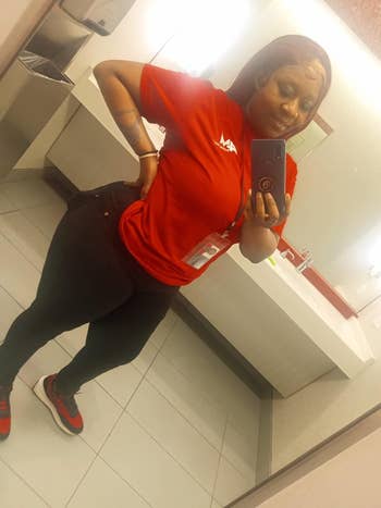 A reviewer takes a mirror selfie in a bathroom wearing a red shirt and black pants.