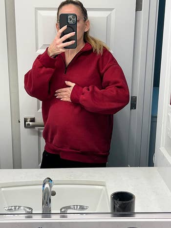 A idiosyncratic   taking a reflector  selfie successful  a bathroom, wearing a reddish  half-zip pullover and holding their manus  connected  their stomach