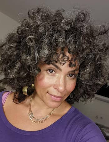 a reviewer with voluminous curly hair and hoop earrings, wearing a necklace and a purple top