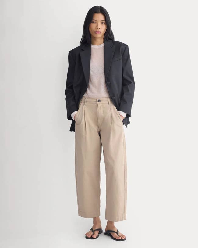 Person in oversized blazer, sheer top, wide-leg trousers, and sandals. They stand facing forward, hands in pockets, on a plain background