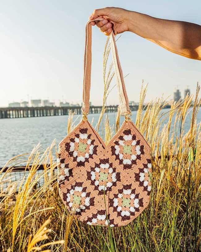 Hand holding a crochet-patterned handbag outdoors near a body of water and grasses