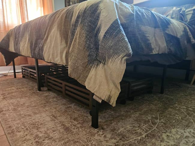 Bed with abstract-patterned comforter and elevated storage underneath, neatly organized with wooden crates. Rug is partially visible