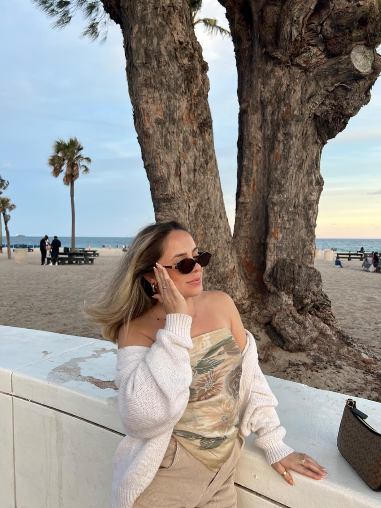 Person connected  a beach, wearing sunglasses and a floral off-shoulder apical  with a airy  sweater, leaning against a railing with thenar  trees successful  the background
