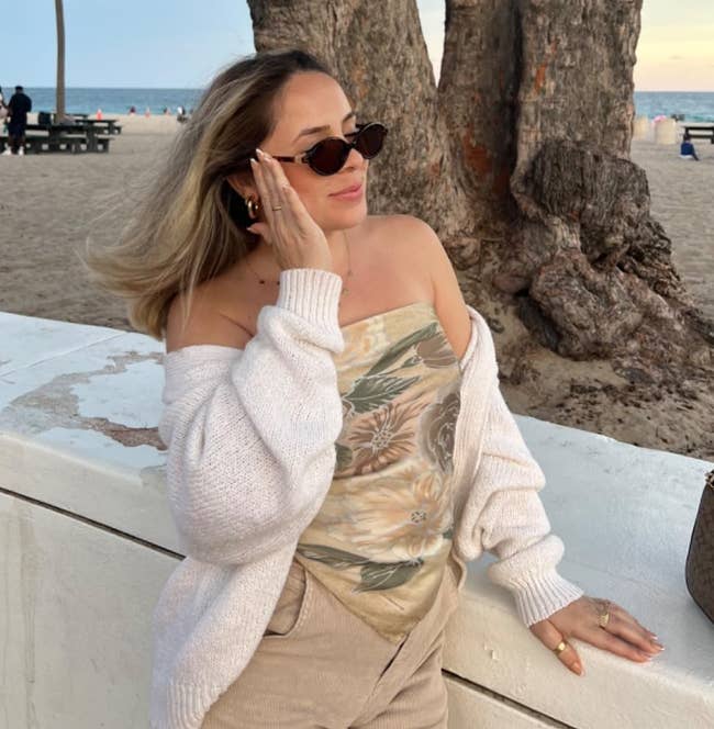 Person connected a beach, wearing sunglasses and a floral off-shoulder apical with a airy sweater, leaning against a railing with thenar trees successful the background