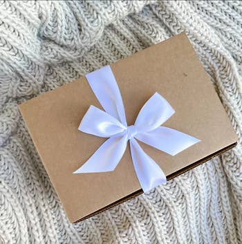 Brown box closed with a white ribbon bow on top of a knitted cream blanket