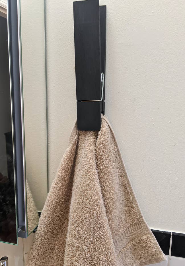 a reviewer photo of a large wall-mounted clothespin mounted on the wall and holding a tan hand towel 