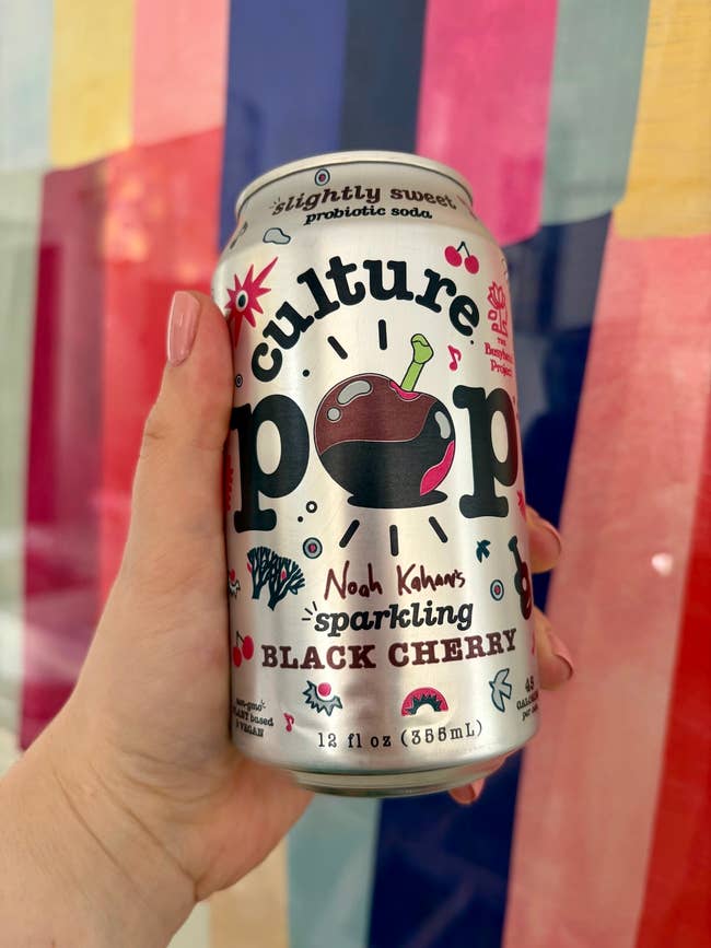 Hand holding a tin  of Culture Pop Noah Kahan's Sparkling Black Cherry soda, with probiotic benefits advertised connected  the can