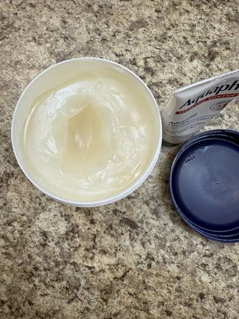 Open jar of Aquaphor healing ointment connected  a countertop, with lid and packaging beside it