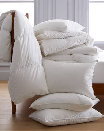 A stack of various sized plain white pillows and comforters set against a simple room background, suggesting comfortable bedding options
