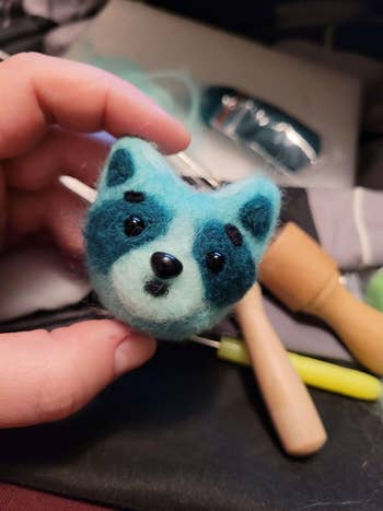 Hand holding a felted blue raccoon face, with crafting tools in the background
