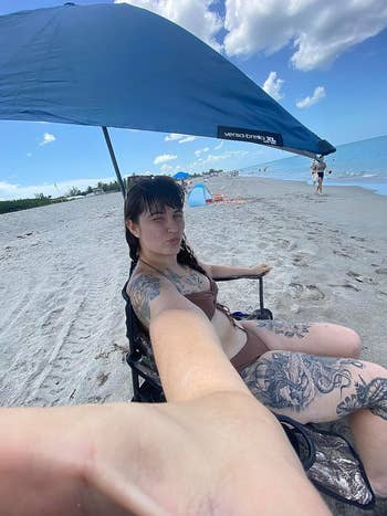 Person at the beach in a wheelchair, taking a selfie under a sun shade. They are wearing a swimsuit