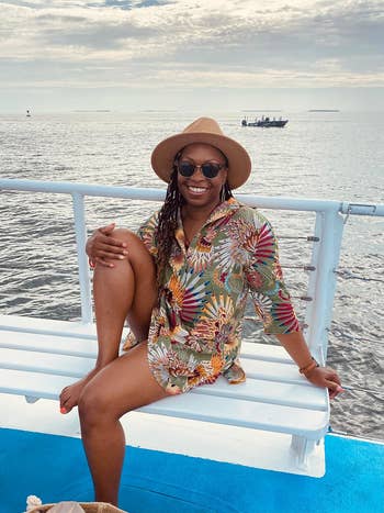 Woman in floral outfit and hat sitting on boat railing with ocean and ship in the background