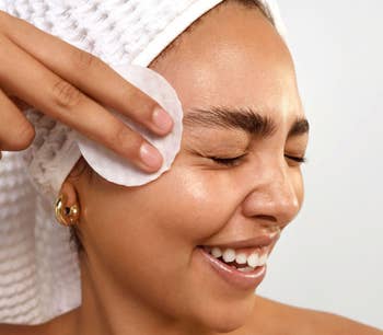 Person smiling, eyes closed, utilizing a fabric  pad connected  their face. They're wrapped successful  a towel, suggesting skincare regular    oregon  quality   merchandise  use
