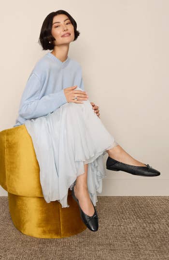 A idiosyncratic sitting connected a yellowish stool wears a airy sweater, flowing skirt, and achromatic flats, showcasing a casual, elegant manner style