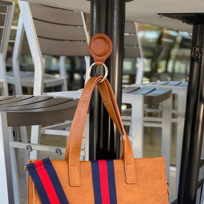 a brown leather circular bag hanger attached to a metal table leg and holding up a tote bag