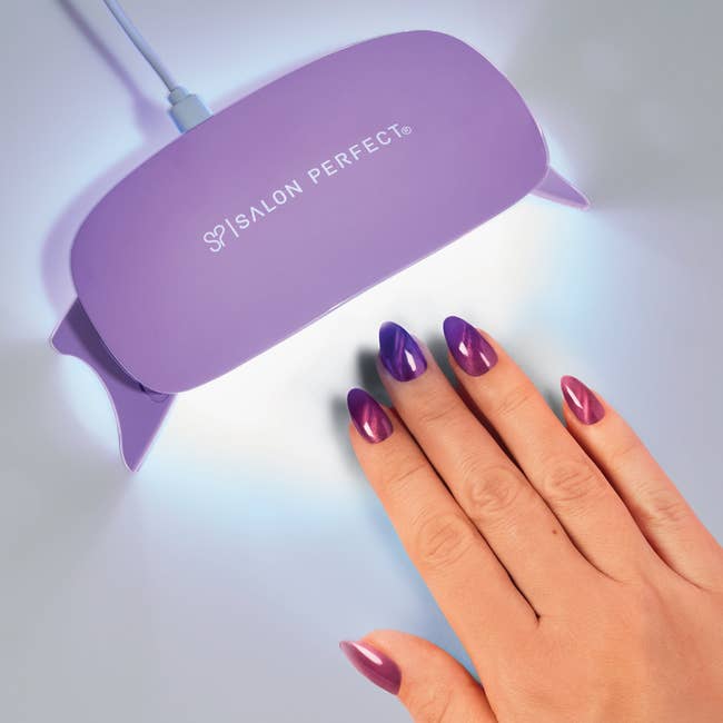 A manus with glossy purple nails nether a UV lamp with the Salon Perfect logo, showcasing a nail polish curing process