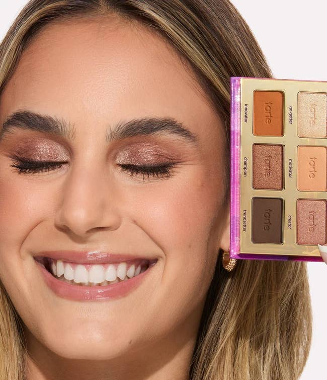 Person smiling with eyes closed, holding a constitution palette adjacent face, showing eyeshadow shades