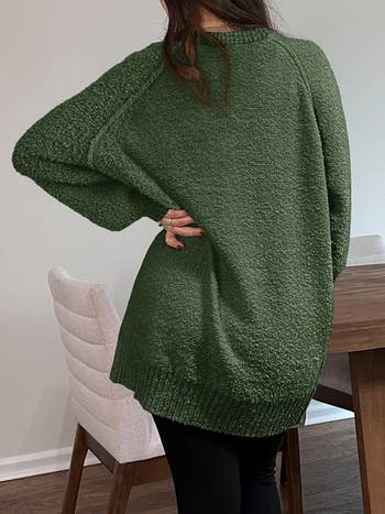 A person is standing with their back to the camera, wearing a textured oversized sweater and black pants, next to a dining table and chairs