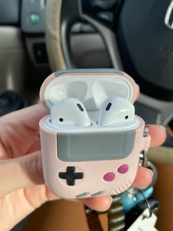 Close-up of a hand holding Apple AirPods in a pink Gameboy-themed case with a retro design. The background shows car interior elements