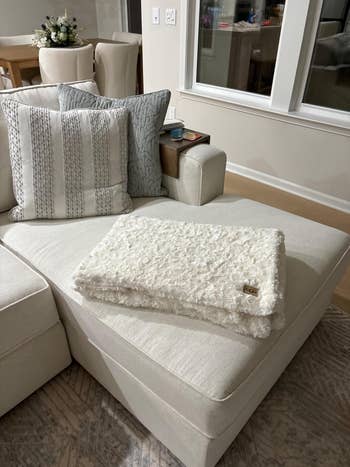 A plush, textured white blanket is neatly folded on a beige sectional sofa with decorative pillows, set in a neutral-toned living room