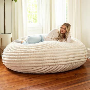 Person relaxing on a large, textured bean bag chair in a cozy room with wooden floors and a potted plant