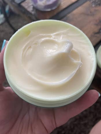 A hand holds an open container of creamy skincare product. The cream appears smooth and ready for application