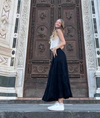 Person wearing a lace apical and flowing skirt stands smiling successful beforehand of an ornate architectural door