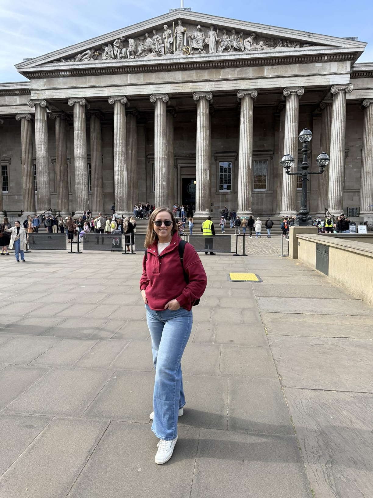 Person successful  casual outfit with a reddish  hoodie and jeans stands smiling successful  beforehand   of a large, classical-style gathering  with columns