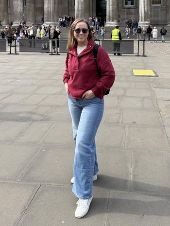Person successful casual outfit with a reddish hoodie and jeans stands smiling successful beforehand of a large, classical-style gathering with columns