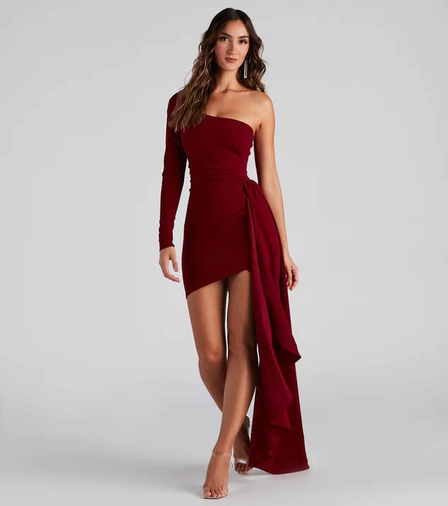 Model wearing burgundy red mini dress with long side train and one long sleeve shoulder, paired with clear open-toed heels