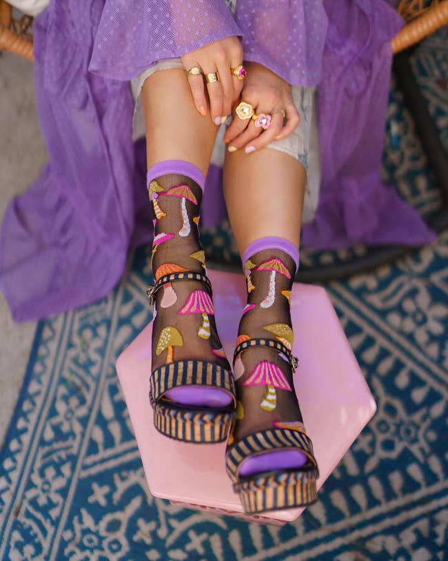 model in black sheer socks with purple ankle and toe trim and large colorful mushrooms peeking out of sandals