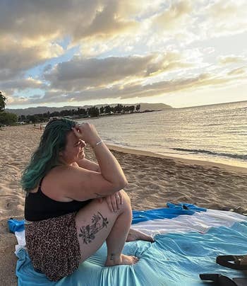 Person sitting connected  a formation  astatine  sunset, looking retired  astatine  the ocean, wearing a achromatic  apical  and leopard-print shorts with a floral tattoo connected  their thigh