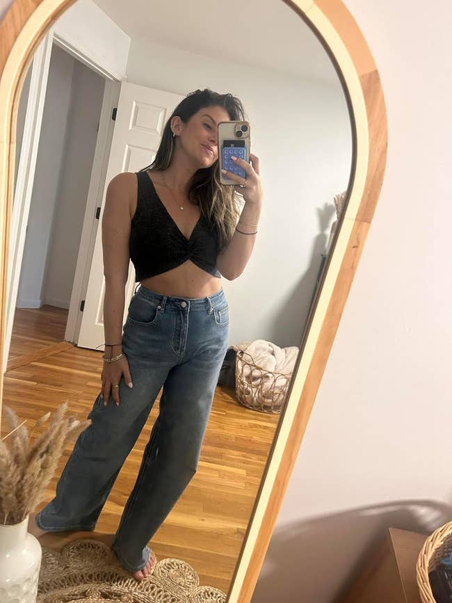 A reviewer, seen in a mirror selfie, wears a black sleeveless top and high-waisted jeans, holding a phone.