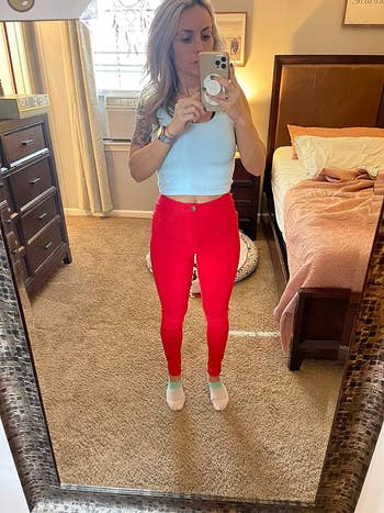 A reviewer taking a mirror selfie wearing a white tank top and red jeans.