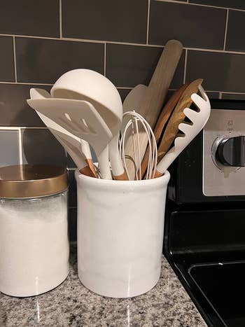 Kitchen counter with a white utensil holder containing spatulas, a whisk, a ladle, and other utensils. Next to it is a jar with a copper lid