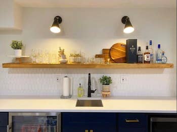 Long light wooden shelf above sink with bottles of alcohol, cutting boards, wine glasses, and plants